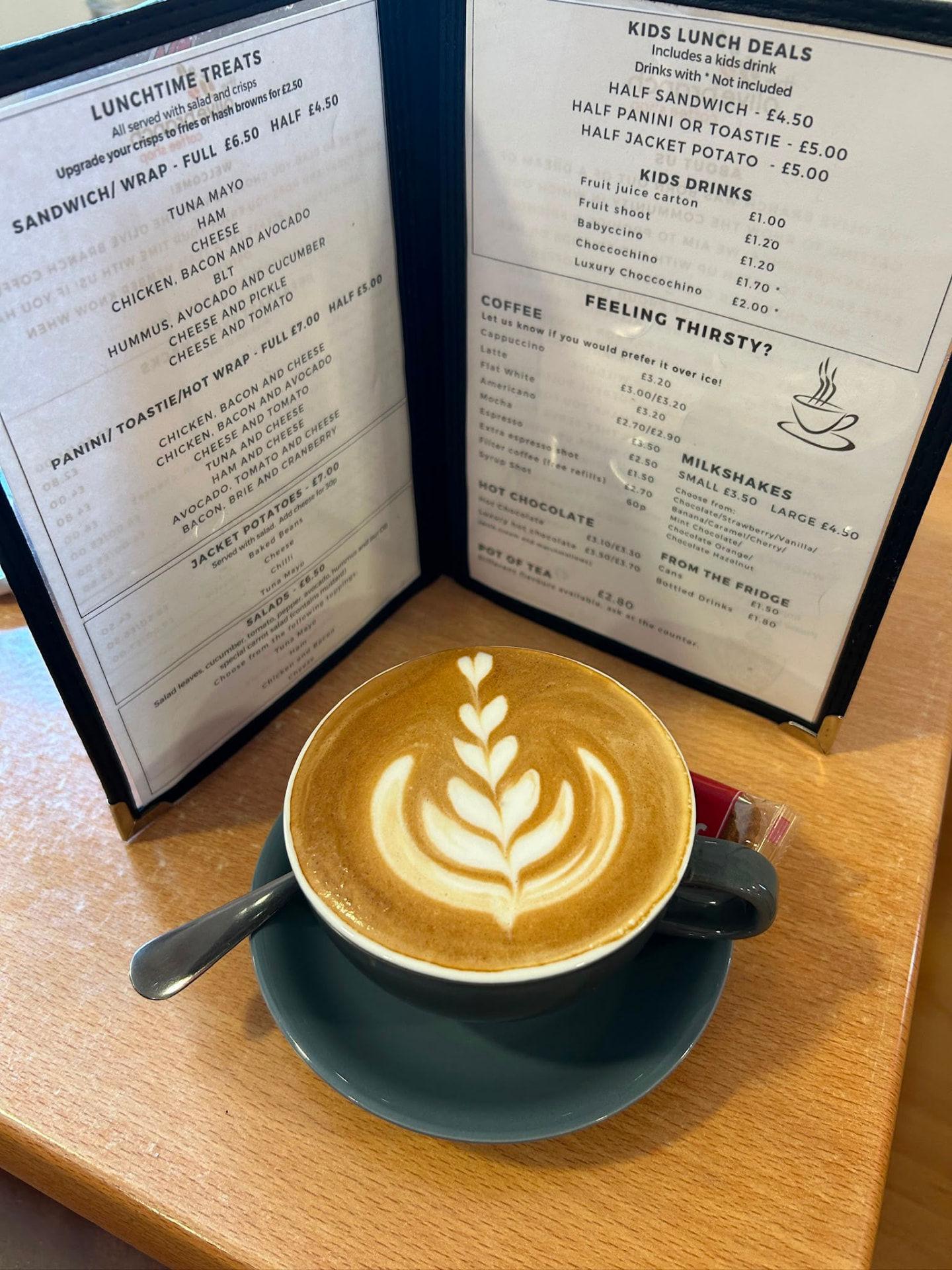 A latte with leaf latte art beside the Olive Branch menu board