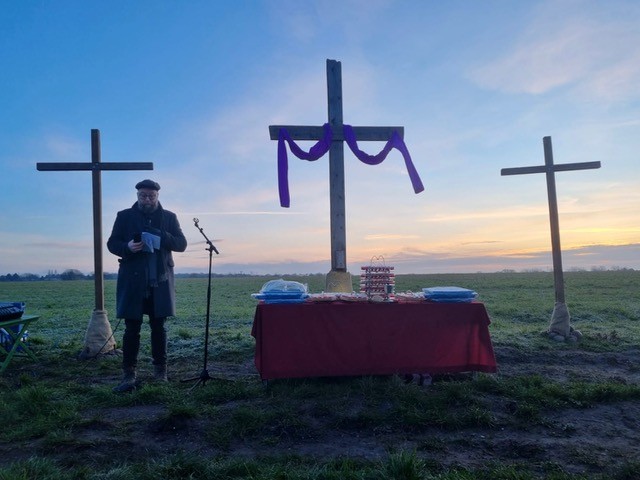 Minister leading an early morning service beside three wooden crosses in a field at sunrise