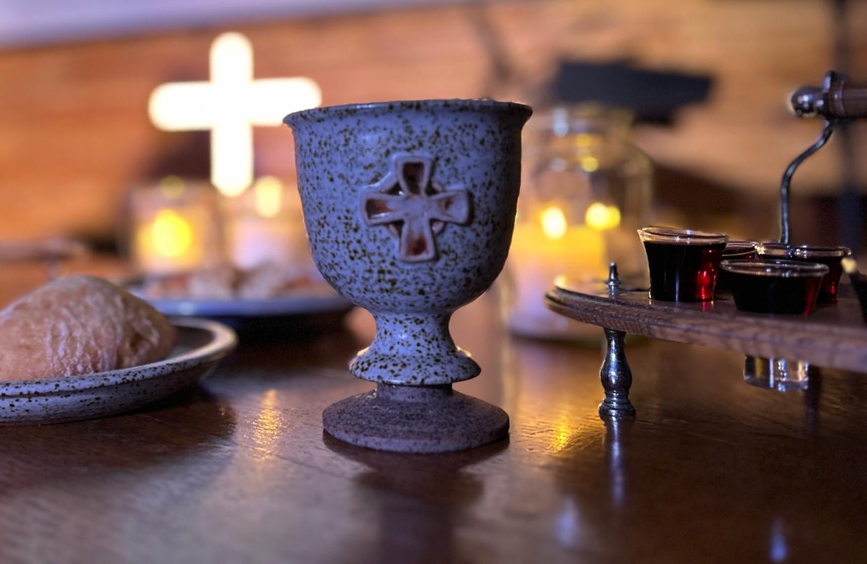 Communion bread and cup on a wooden table with a glowing cross behind