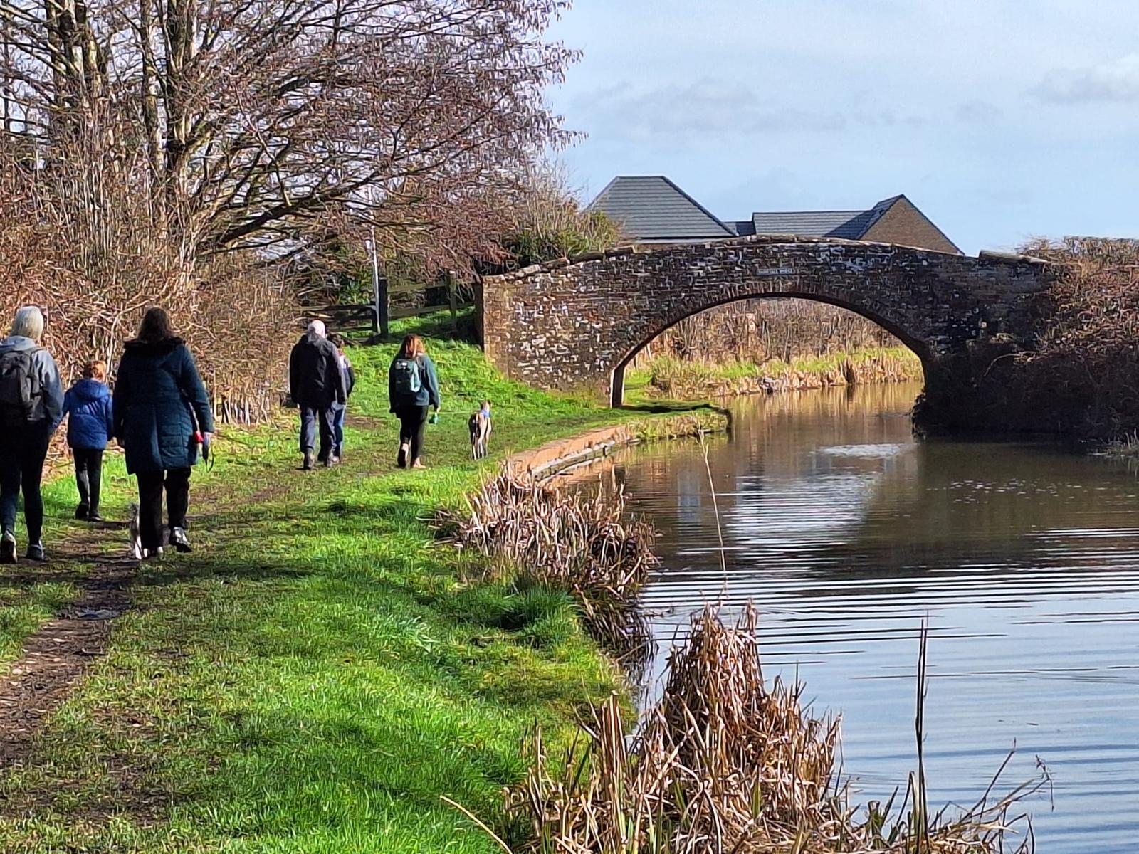 Church walking group following a sunny canal towpath towards a stone bridge
