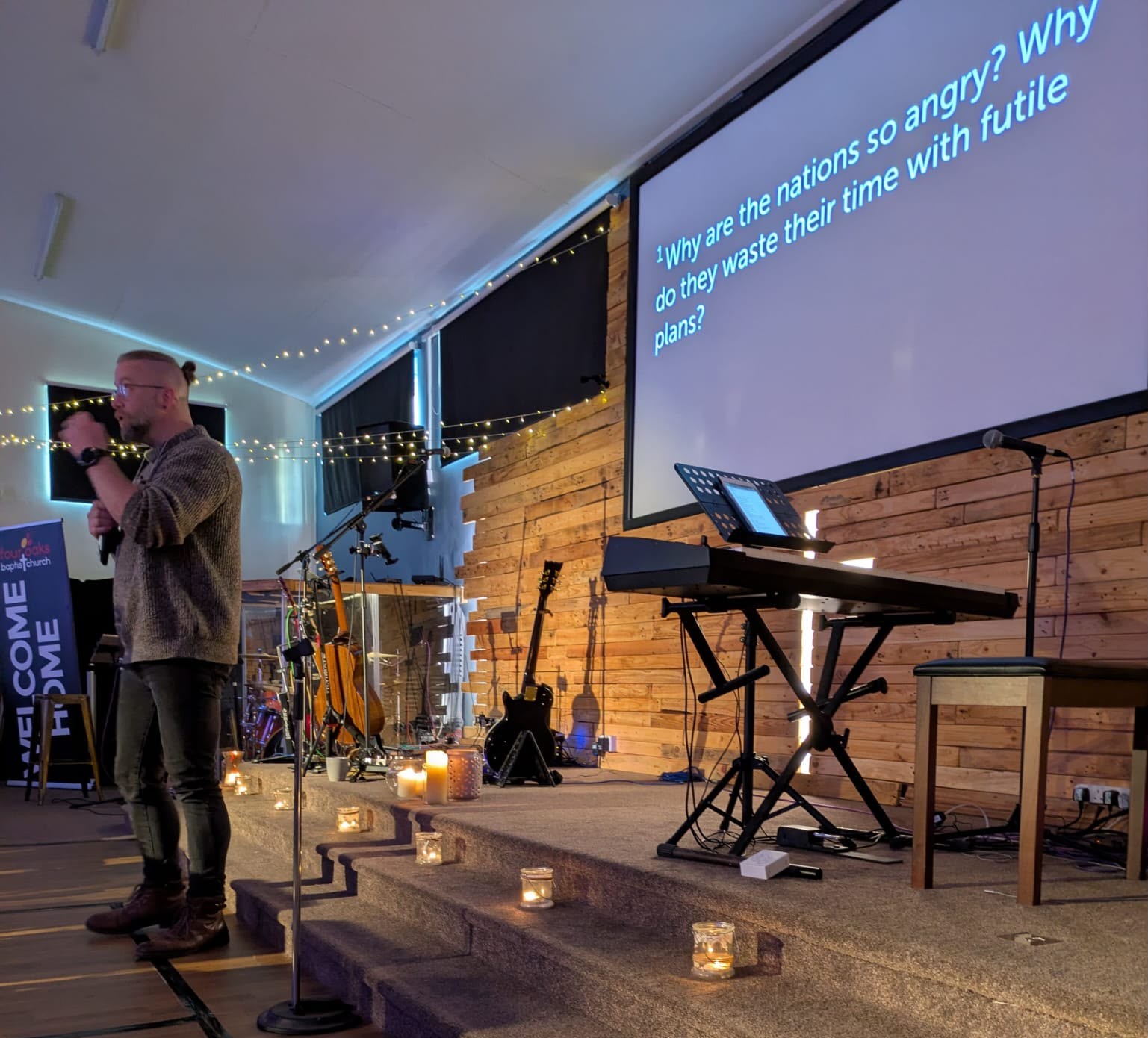 Rev Phil speaking on stage in the evening, candles on the steps, fairy lights and reclaimed wood behind