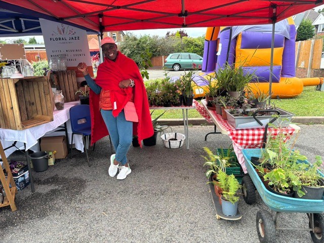 Volunteer wrapped in a red blanket beside plant and craft stalls at the summer fete