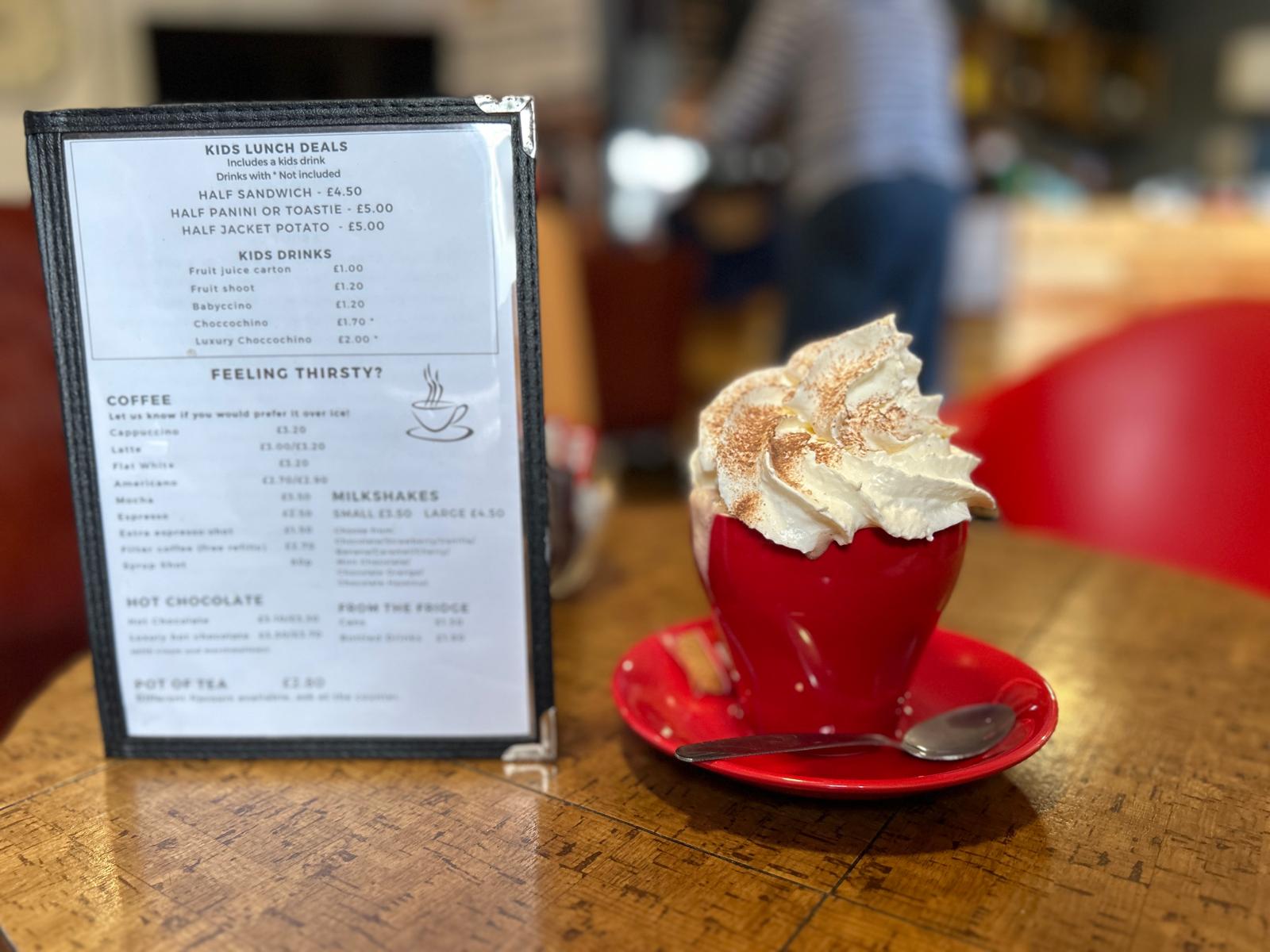 A luxury hot chocolate with cream in a red cup beside the coffee shop menu