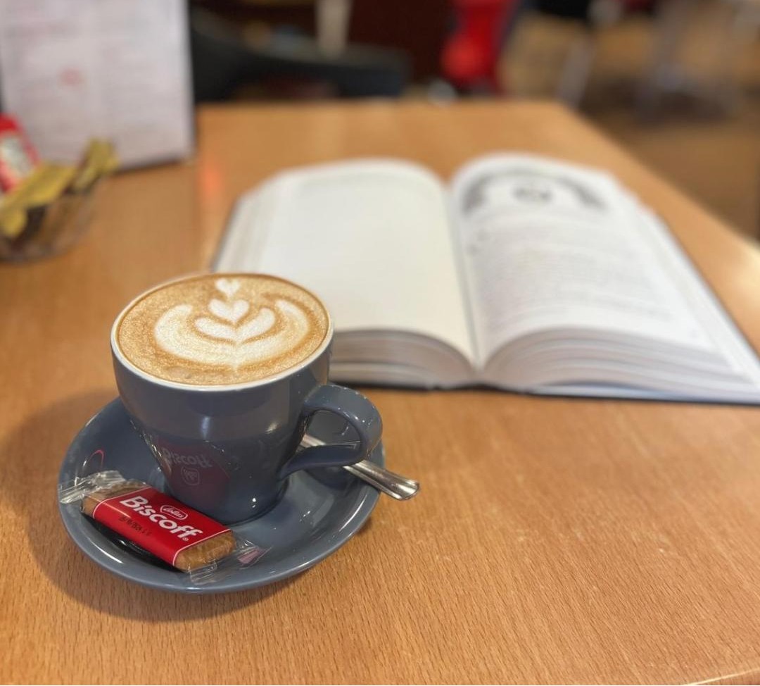A latte with heart-shaped latte art and a Biscoff biscuit beside an open book in the Olive Branch coffee shop