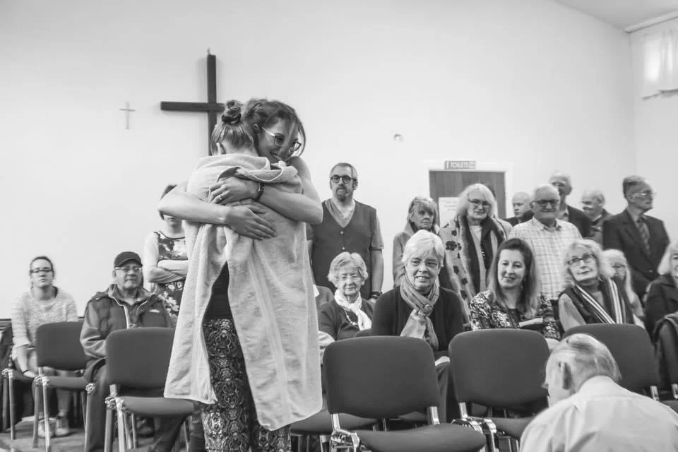 Two people embracing after a baptism, wrapped in a towel, as the congregation looks on