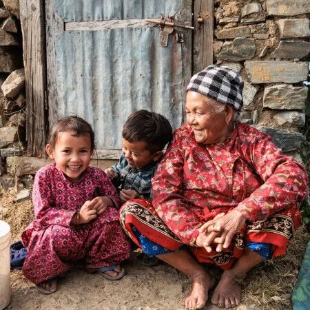 A grandmother and two young children laughing together outside their home in a rural village
